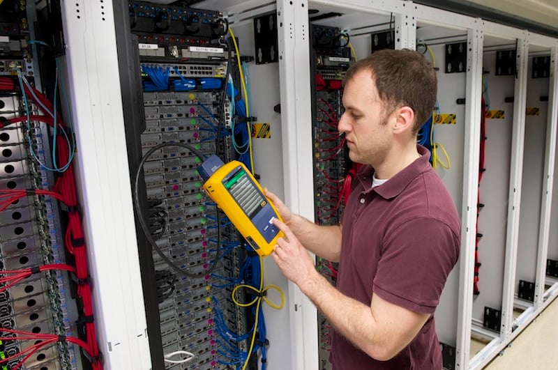 Man standing in front of data server rack, holding a yellow and gray testing device in his hand