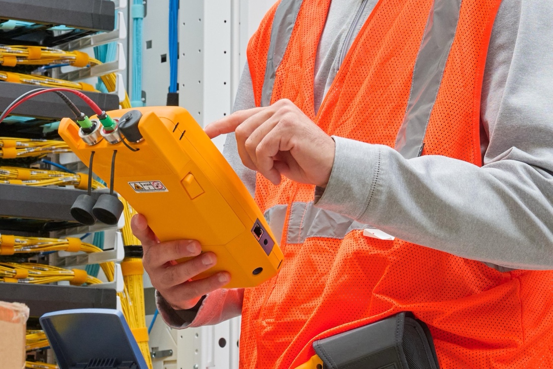 Man in safety vest using a yellow handheld device as he stands in front of a server rack.