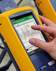 A technician's hand pinching and zooming in on a device screen showing an OTDR trace.