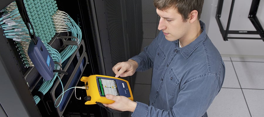 Man in blue shirt using a yellow handheld device as he stands in front of a server rack