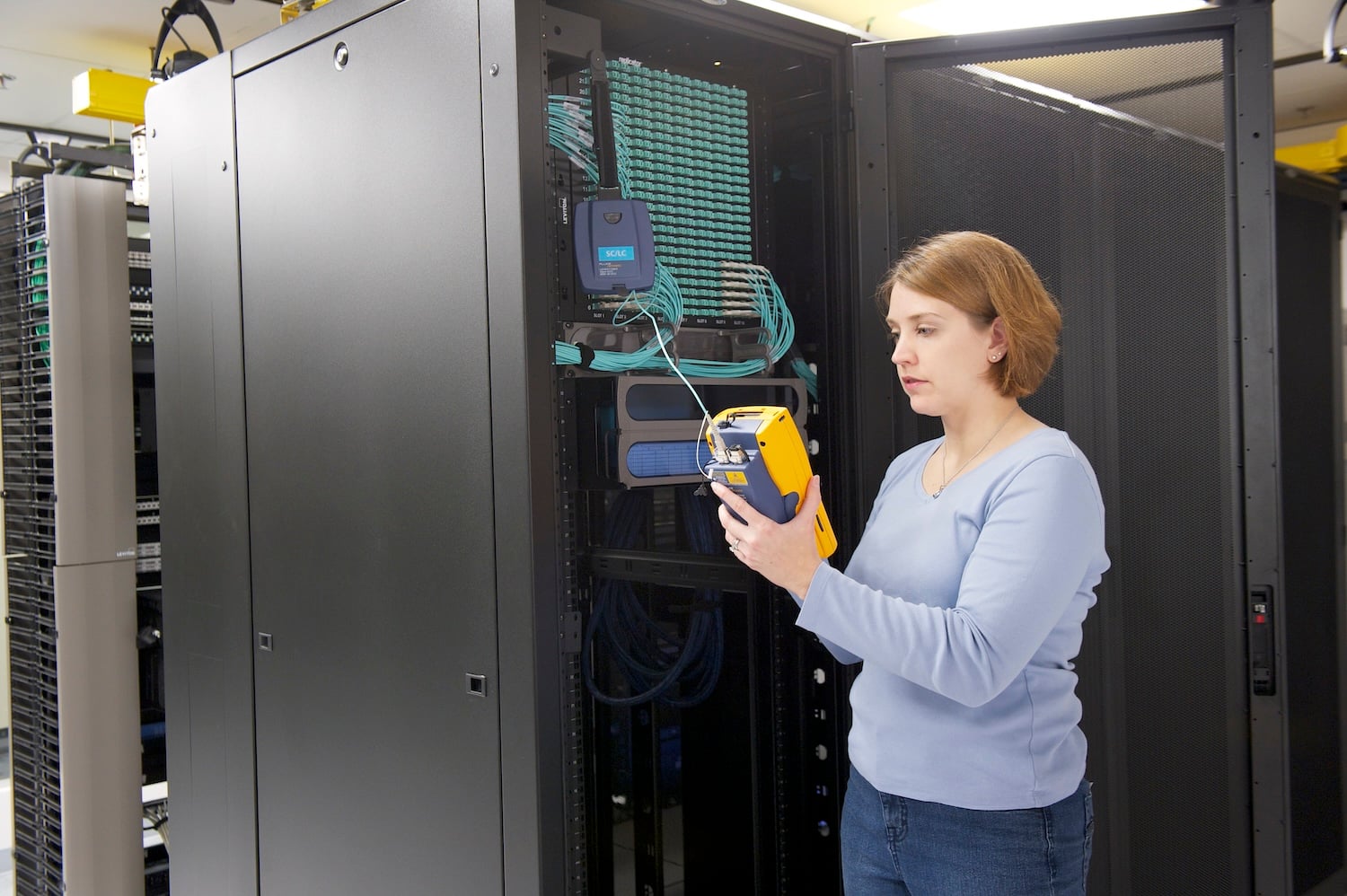 Woman stands in a server aisle in a data center, holding a yellow instrument she's using to test fiber cable links.